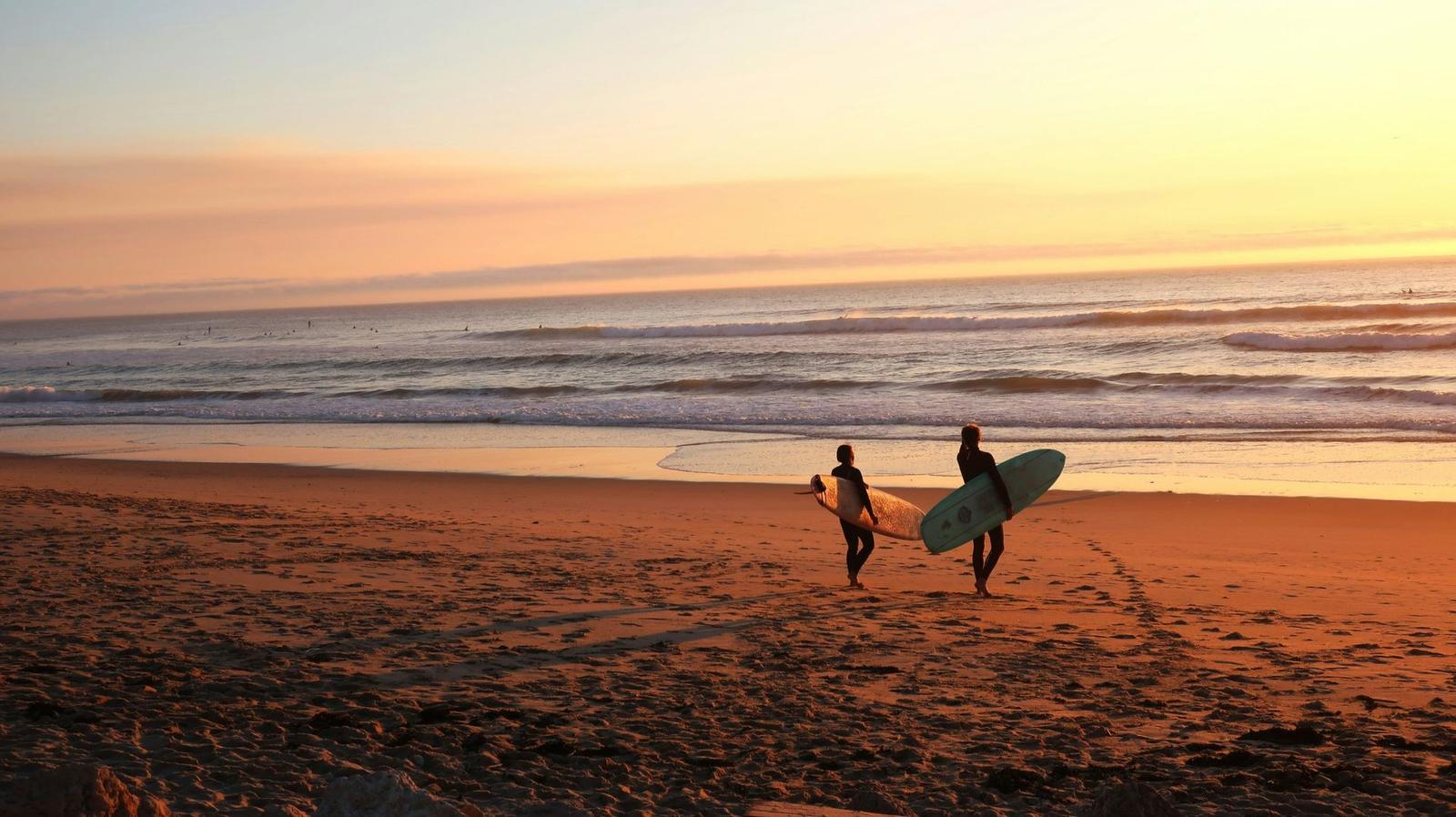 Two surfers walking along the beach at sunset with boards under their arms